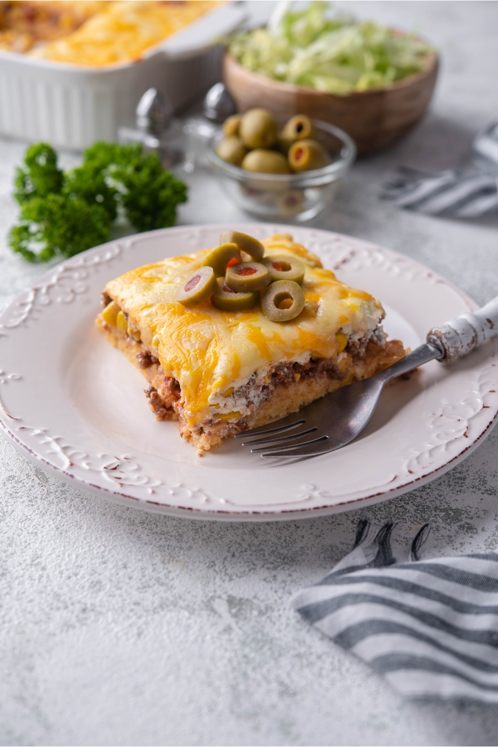 A serving of cornbread taco casserole on a white plate with a garnish if sliced green olives and a fork is on the plate.