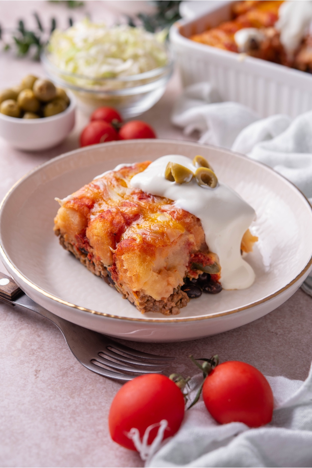 A square serving of taco tater tot casserole with sour cream dripping down the side of the casserole and three slices of green olives garnished on top.