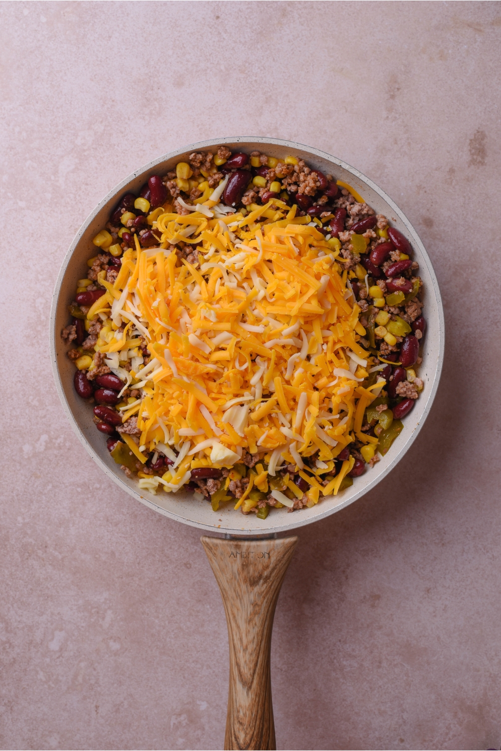 A grey skillet filled with cooked ground beef mixed with black beans, jalapeños, corn, and a pile of shredded cheese on top.