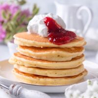 A stack of five flapjack pancakes on a plate with whipped cream and jam on top.