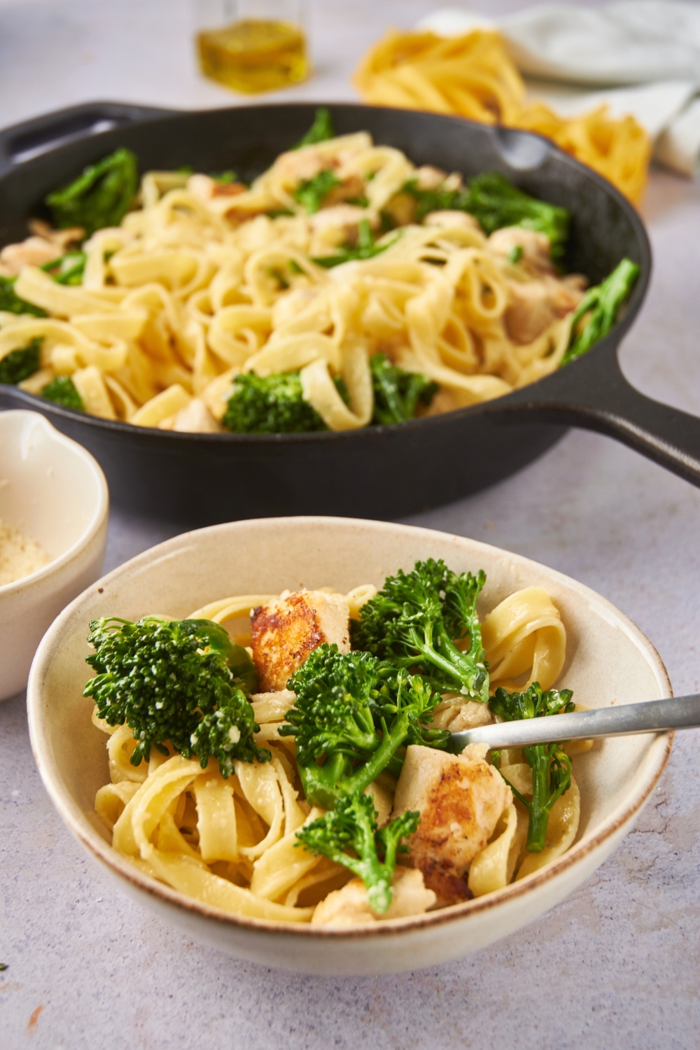 A bowl of broccoli chicken alfredo in front of a skillet filled with more of the pasta.