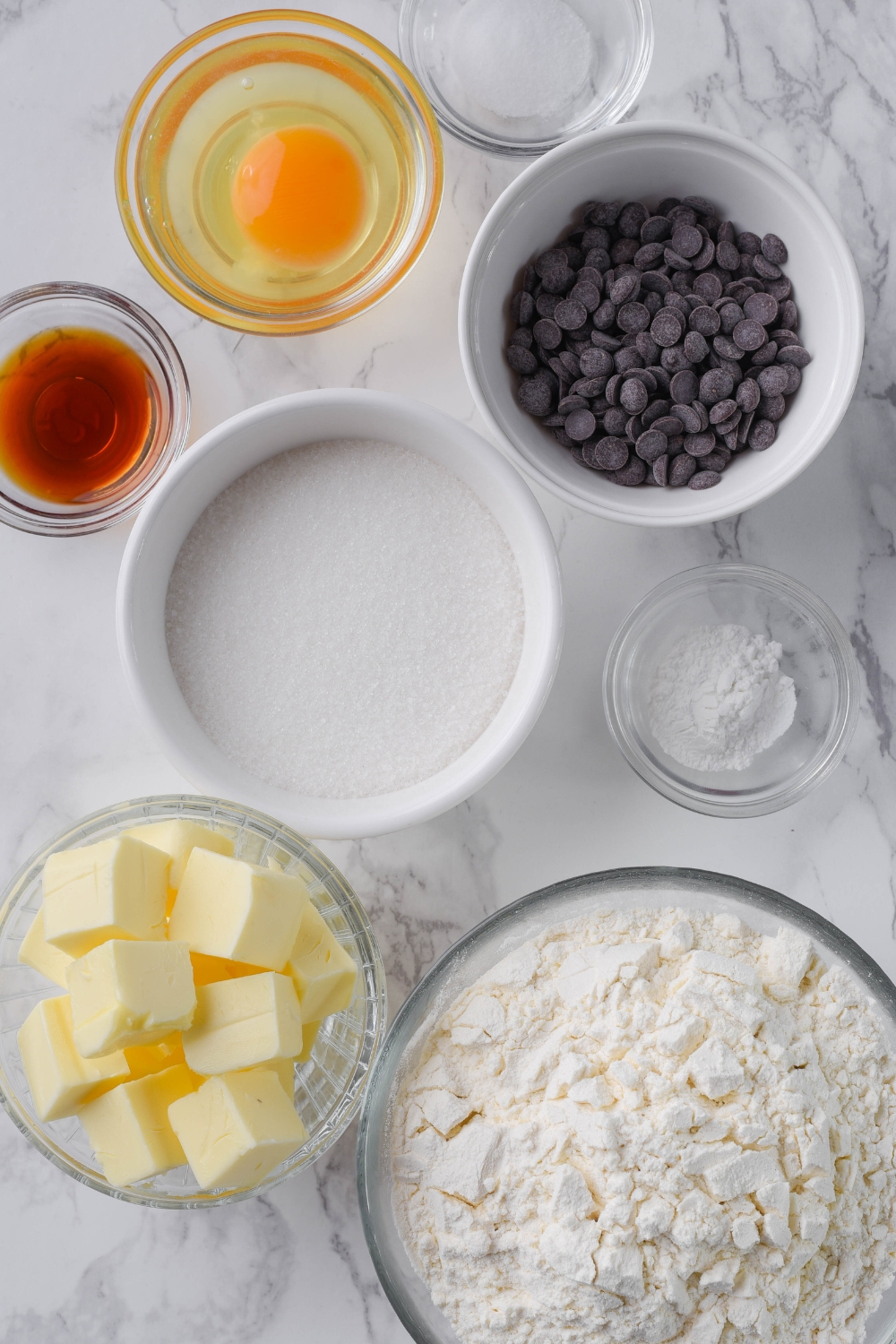 A countertop with multiple bowls with ingredients like flour, butter, sugar, baking powder, vanilla extract, egg, salt, and chocolate chips.