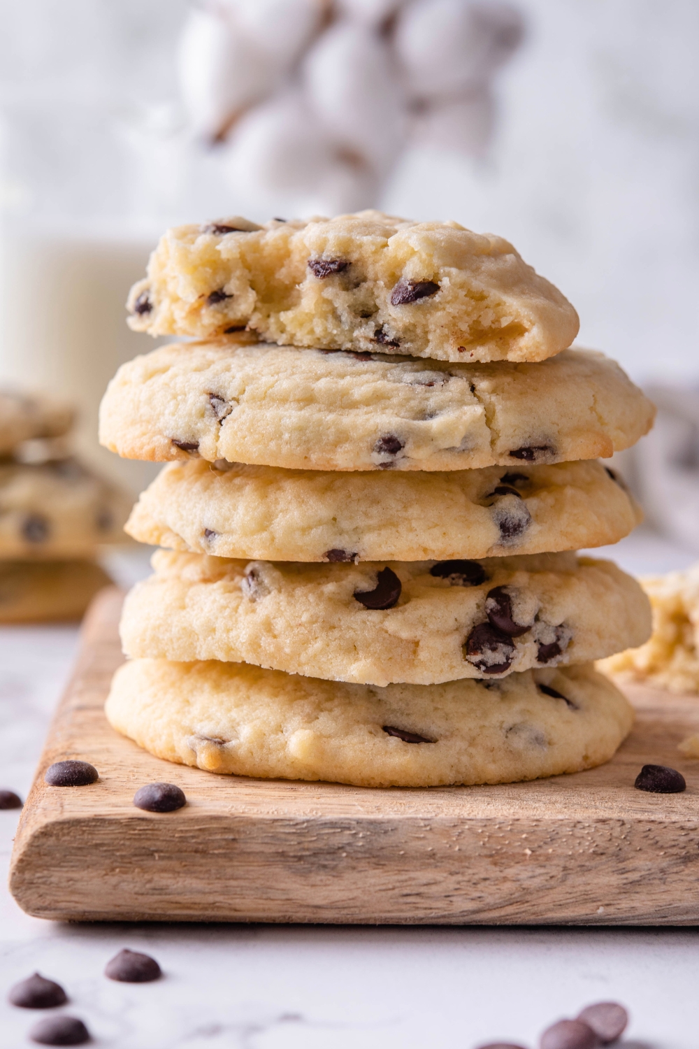 A stack of six chocolate chip cookies on a wooden board. The top cookie has a bite out of the front of it.
