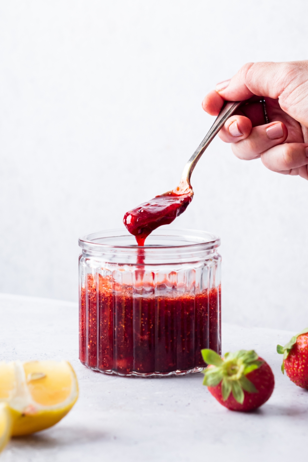 A hand holding a spoon with strawberry compote on it over a glass jar of the compote.