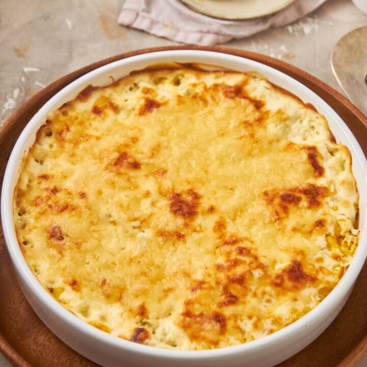 A corn casserole with cream cheese with melted cheese on top in a white bowl on a wooden serving tray.