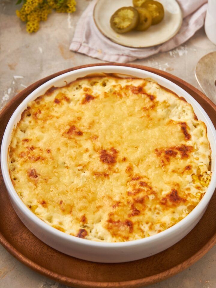 A corn casserole with cream cheese with melted cheese on top in a white bowl on a wooden serving tray.