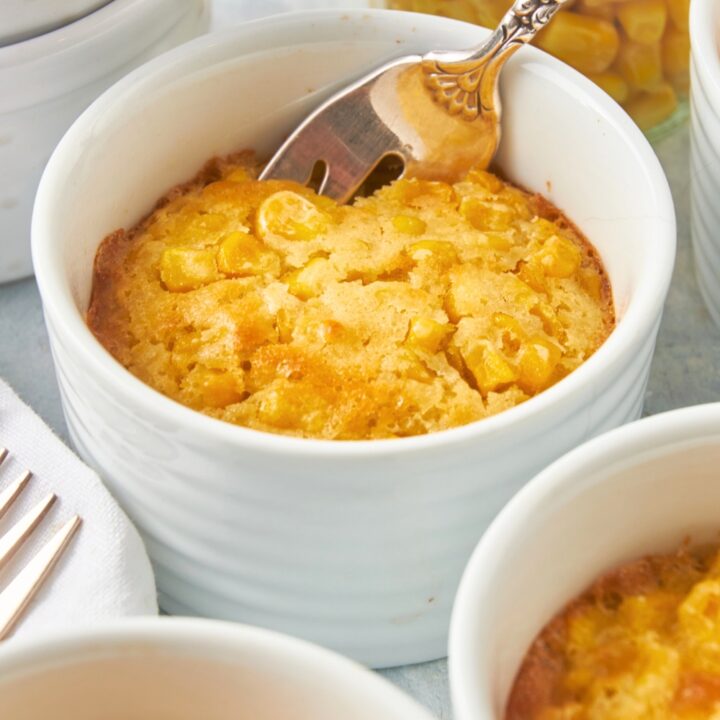 A white ramekin with corn casserole in it with a spoon submerged in the casserole.