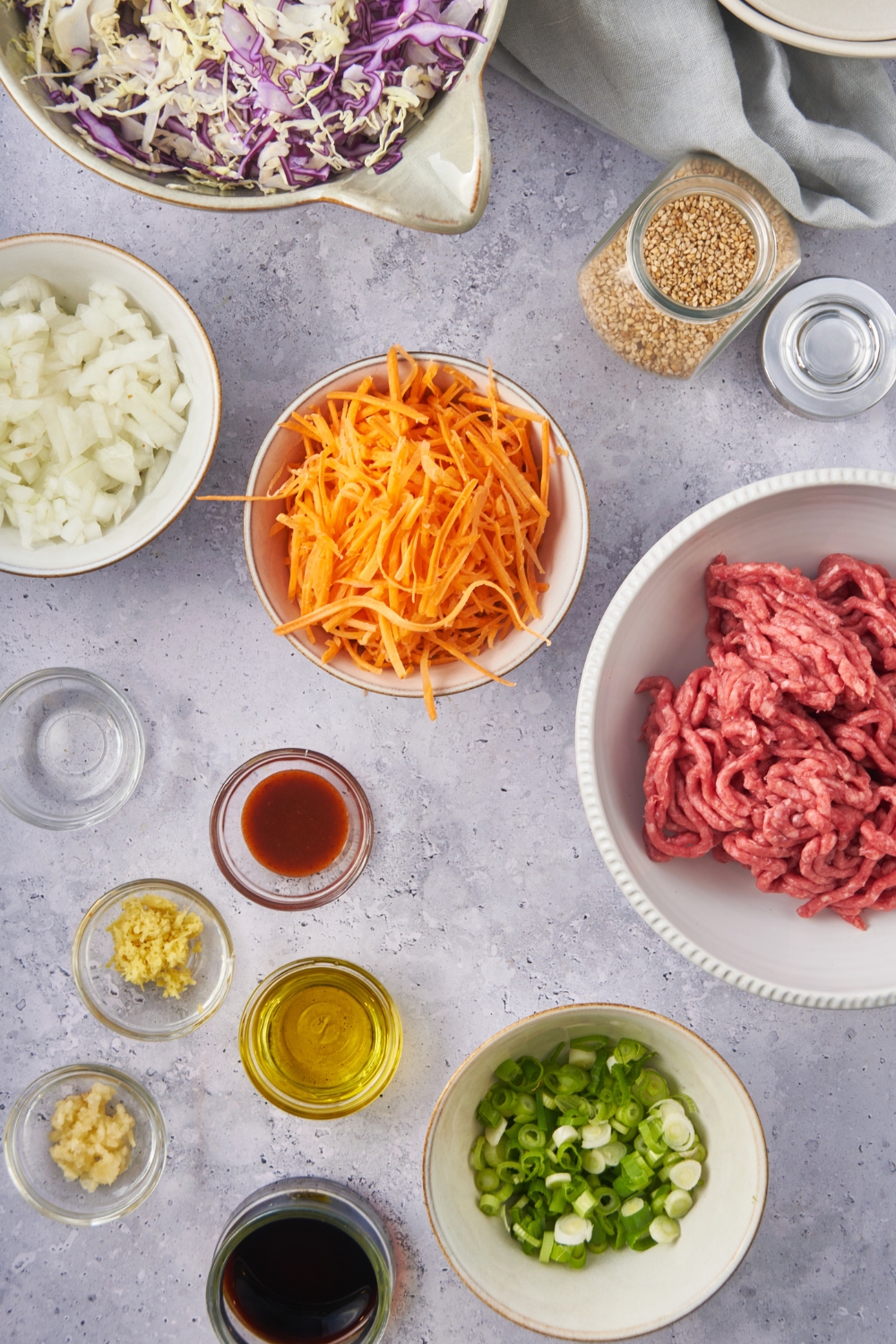 A countertop with multiple bowls containing carrots, ground beef, chopped green onions, olive oil, soy sauce, minced garlic, minced onion, minced ginger, sriracha sauce, and a bowl of coleslaw mix.