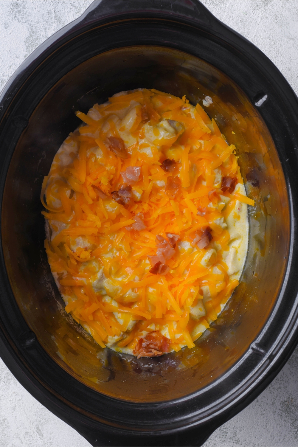 Shredded cheddar cheese and bacon on top of a creamy chicken casserole in a crock pot.