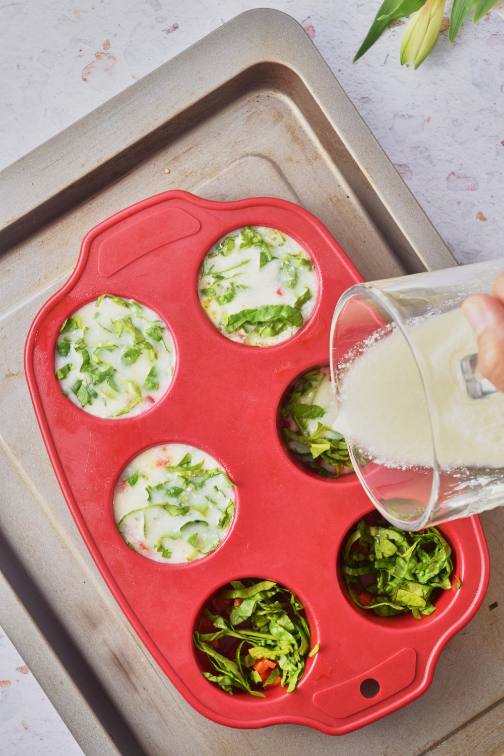 A baking sheet with a silicone muffin mold. A pitcher is pouring the egg mixture overtop the red peppers and spinach.