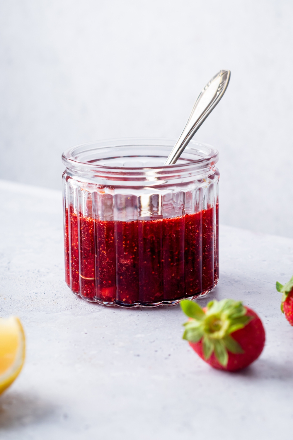 A glass jar that is filled with strawberry compote with a spoon in it.