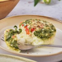 An egg white bite with spinach and red pepper with a piece cut from the front of it. There's a fork in front of the egg bite with a piece of the egg on it.