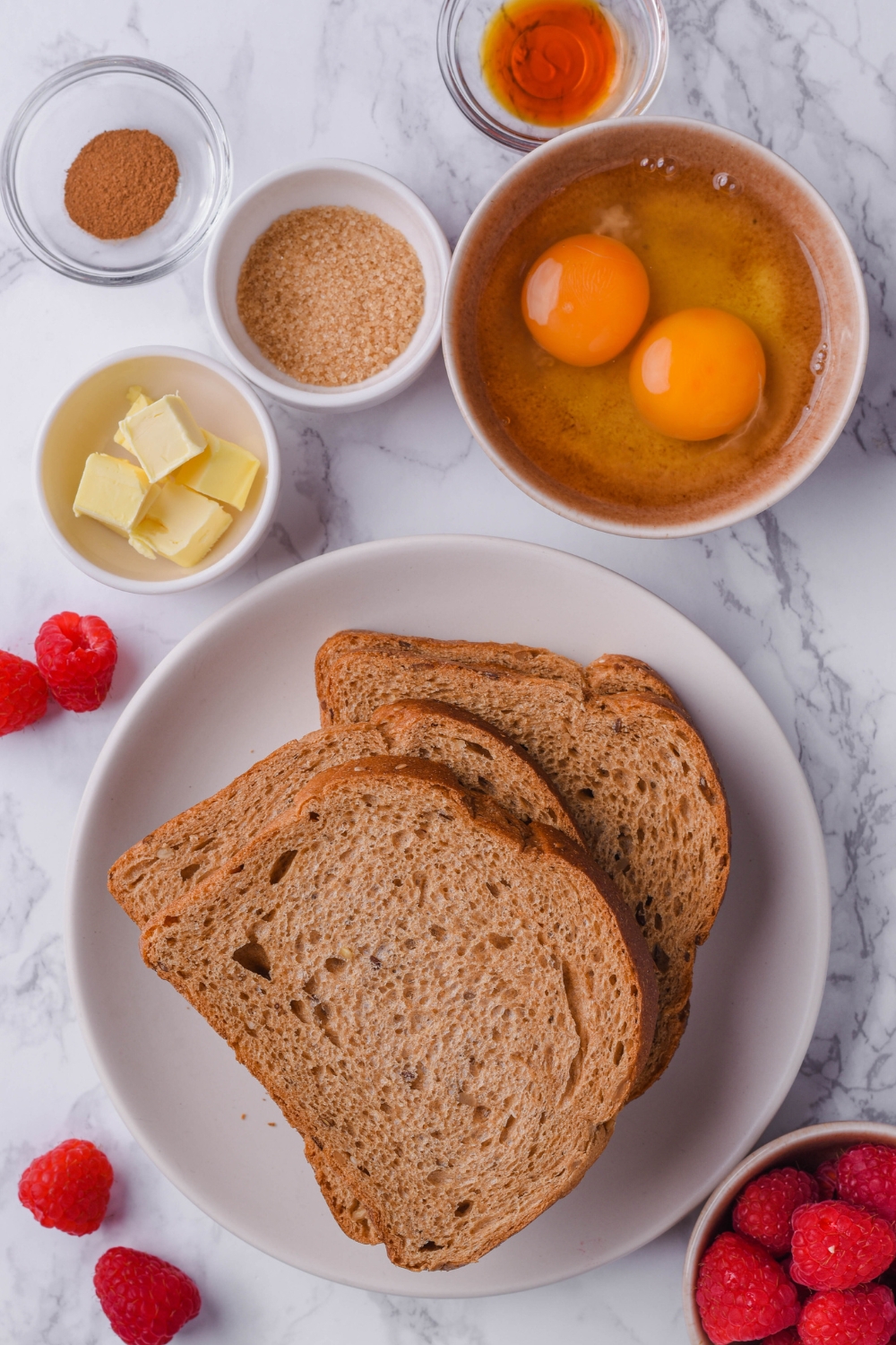A counter top with a bowl with sliced bread, two cracked eggs, butter, brown sugar, cinnamon, and vanilla extract.