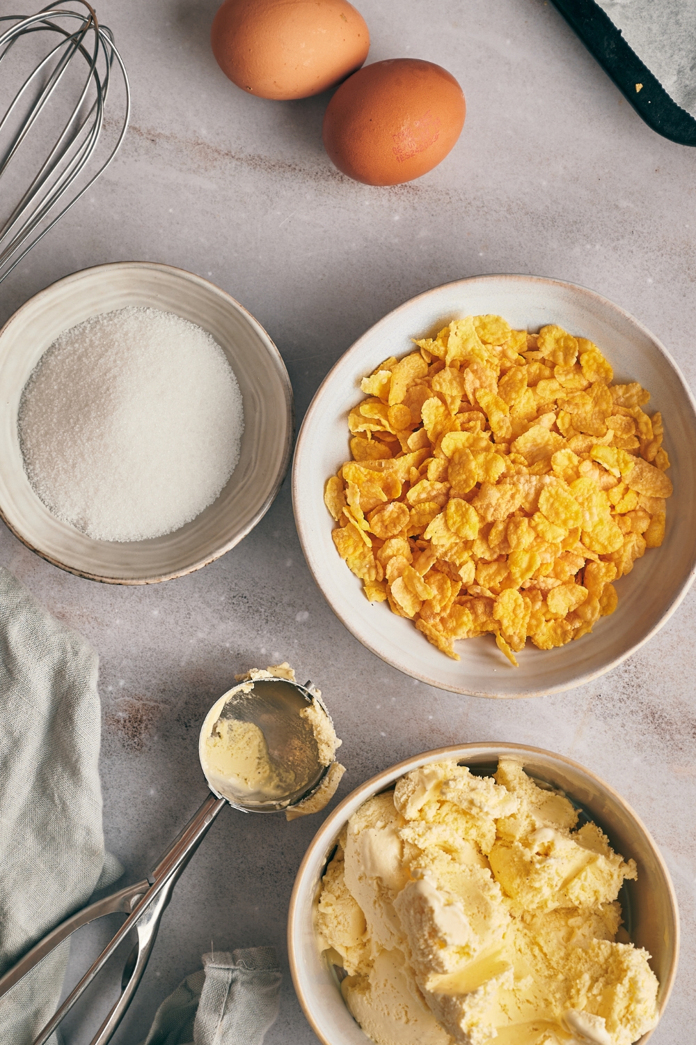A countertop with two eggs, a bowl of sugar, a bowl of cornflakes, and a bowl of vanilla ice cream.