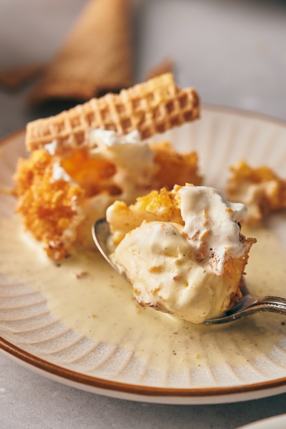 A plate with fried ice cream topped with whipped topping and an ice cream cone. A spoon has a scoop of the melted ice cream and crunchy cornflake topping.