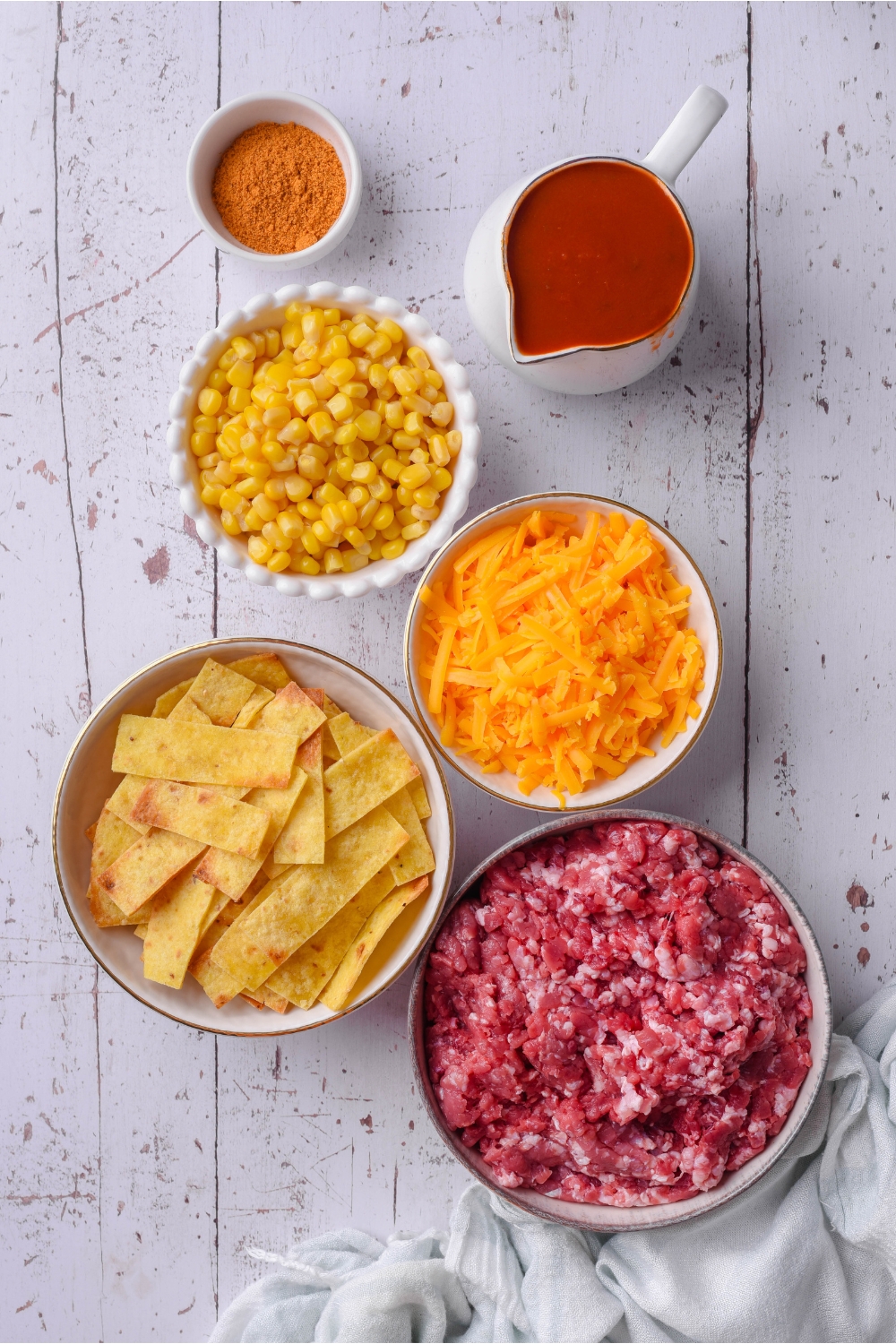 A bowl of corn, a bowl of taco seasoning, a bowl of shredded cheddar cheese, a bowl of fritos, and a bowl of ground beef all on a white counter.