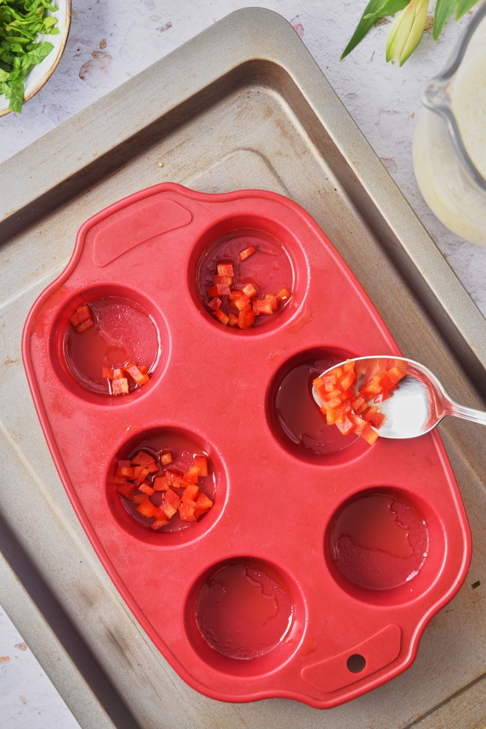 A baking sheet with a silicone muffin mold. A spoon is adding diced red peppers into each mold.
