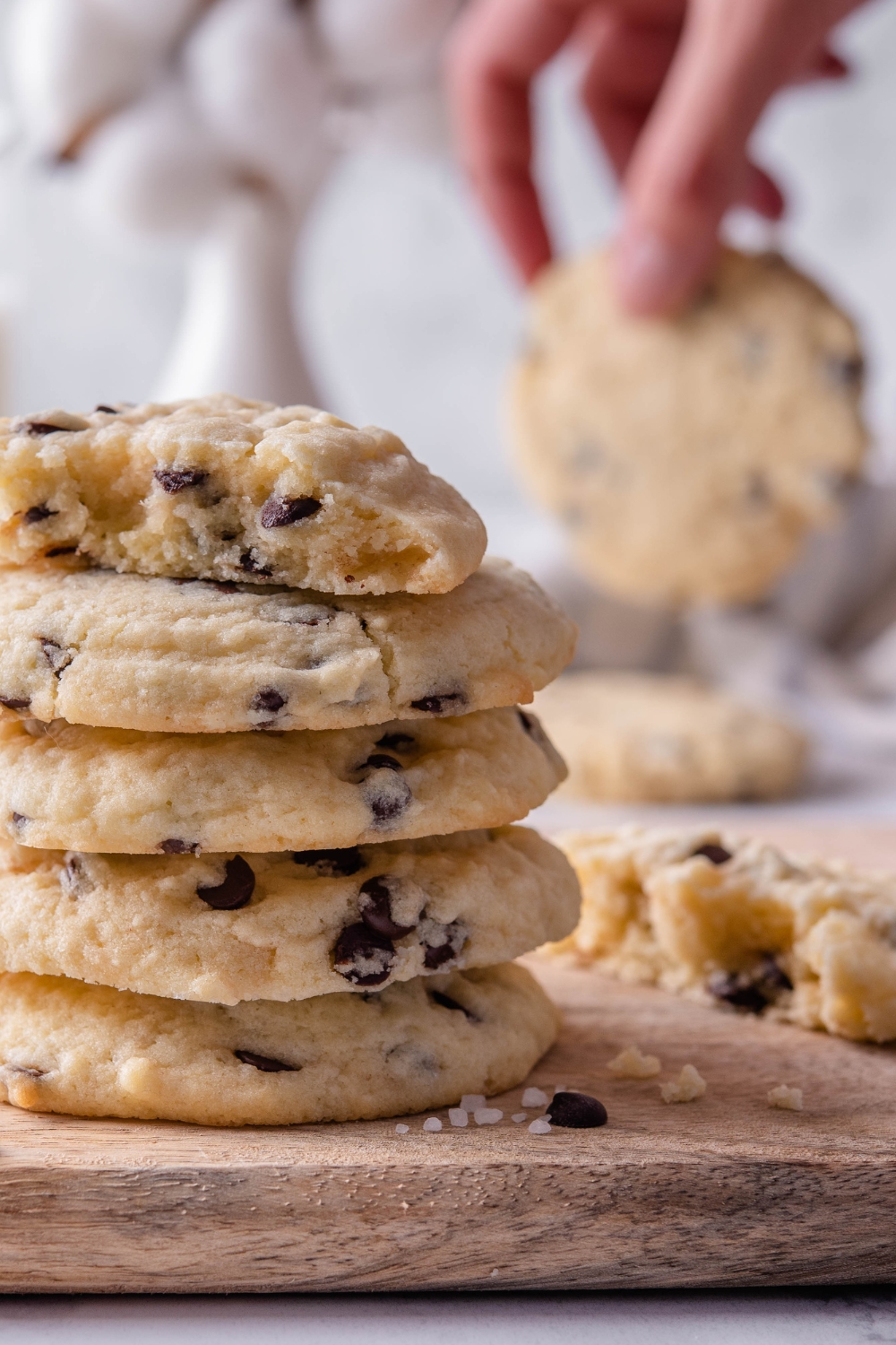 A stack of chocolate chip cookies with one cookie split in half on top. A hand is holding another chocolate chip cookie is in the background.