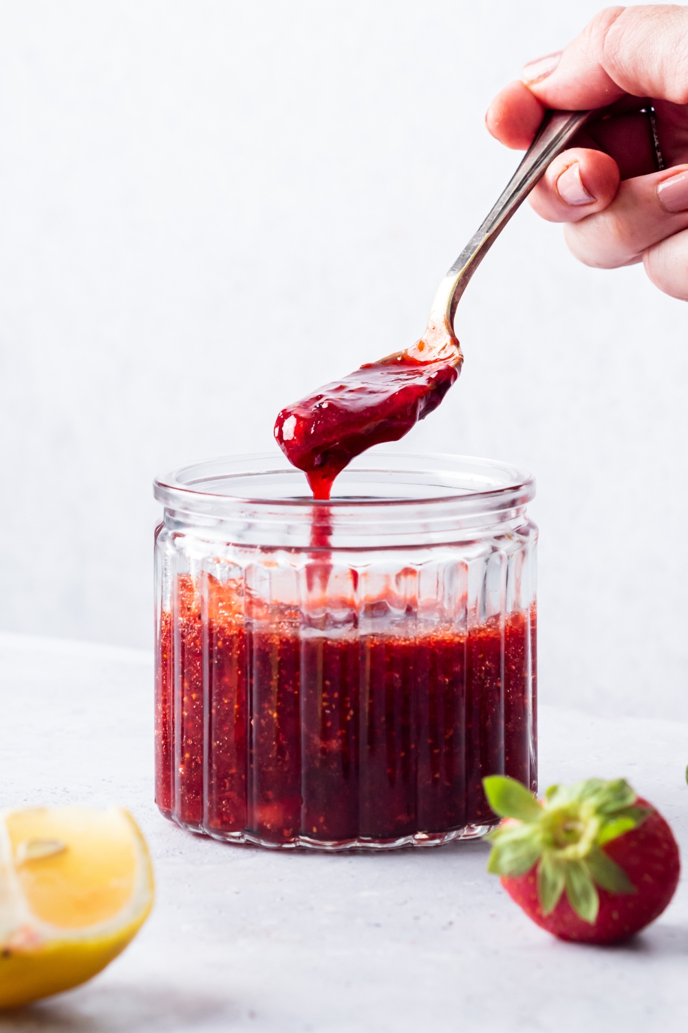 A hand with a spoon that has strawberry compote on it over a jar with the compote.