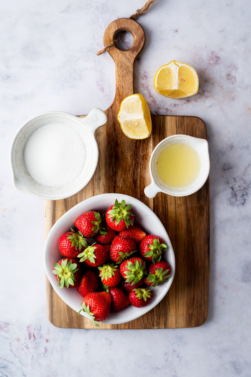 A bowl of strawberries, a bowl of lemon juice, a bowl of sugar, and two lemon wedges on a wood cutting board.