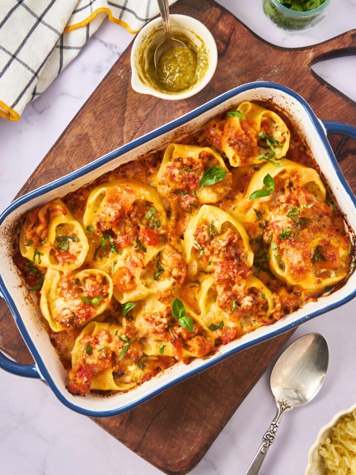 Three rows of four stuffed shells with meat sauce in a baking dish on top of a wooden board on a grey counter.