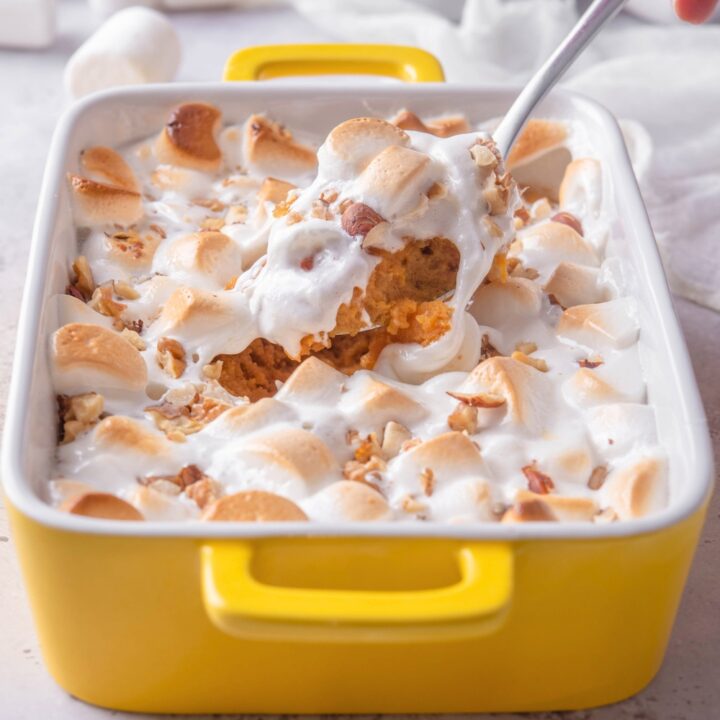 A baking dish filled with sweet potato casserole with canned yams with marshmallows on top. A spoon is scooping some of the casserole out of the dish.