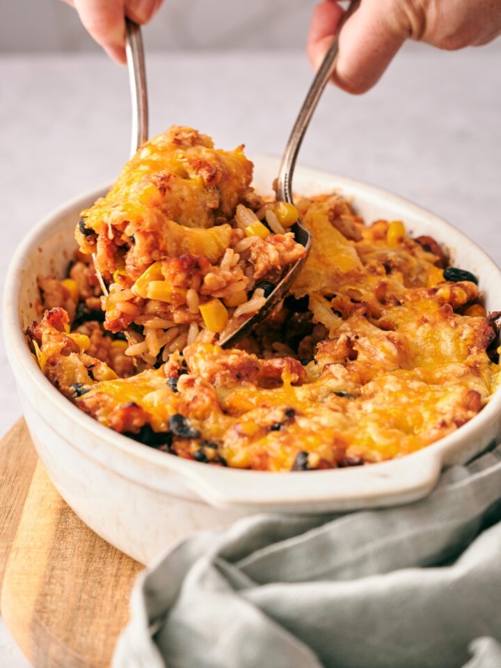 Two hands scooping taco casserole with rice out of a casserole dish filled with the casserole.