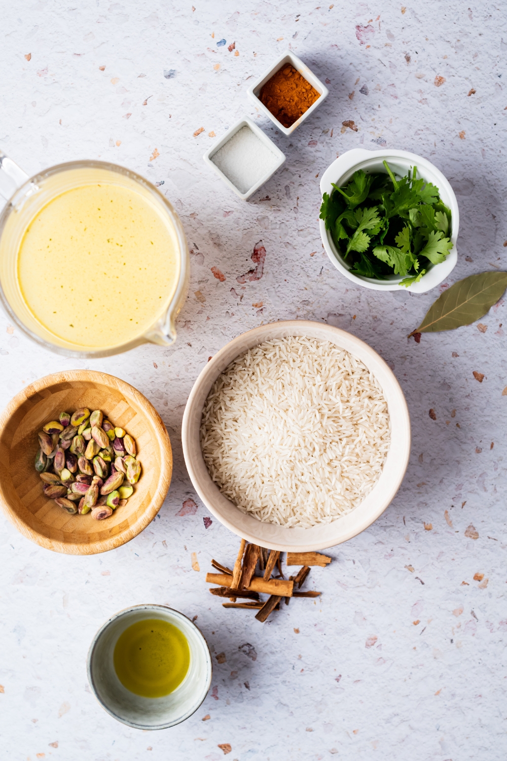 A bowl of rice, a bowl of pistachios, a bowl of broth, a bowl of oil, a bowl of basil, and a bowl of cinnamon on a white counter.