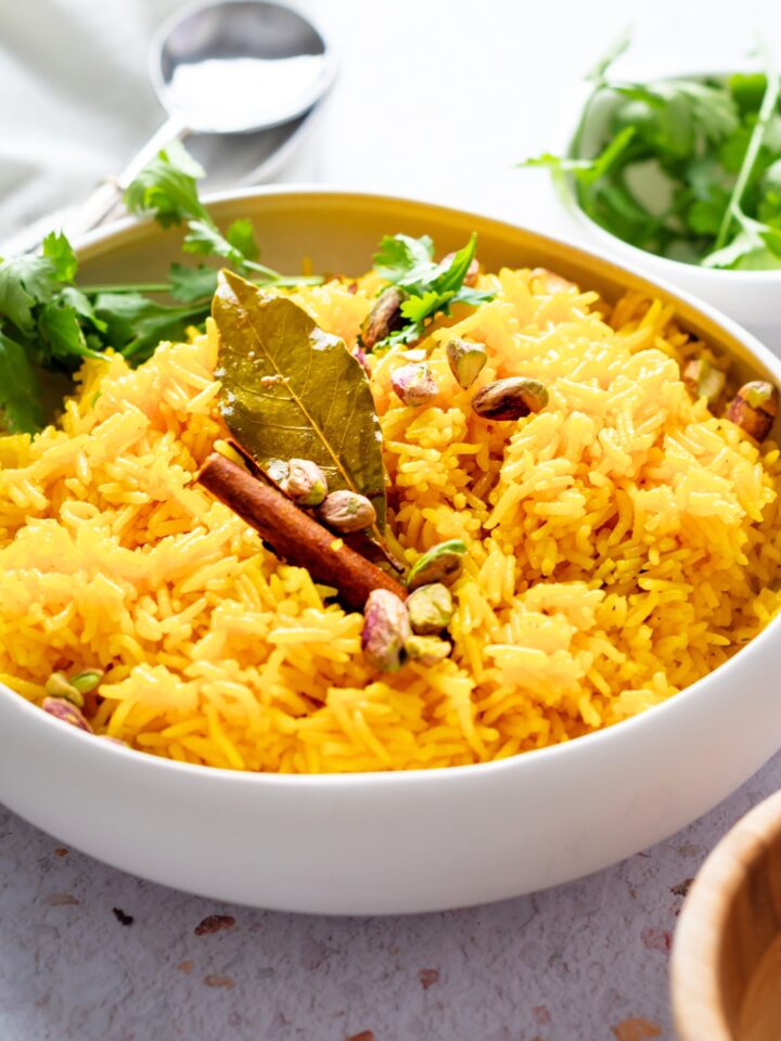 Part of a white bowl filled with turmeric rice with a bay leaf and cinnamon stick on top.