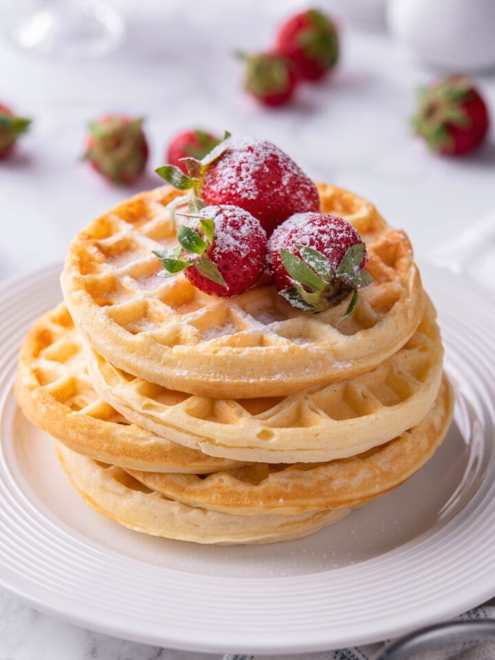 Three strawberries on a stack of five waffles on a white plate.