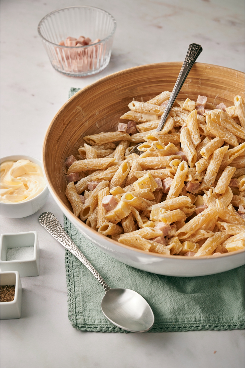 A brown and white bowl with penne pasta, cubed ham, and cubed cheese tossed in a creamy dressing, with a spoon in the bowl and a spoon next to the bowl.