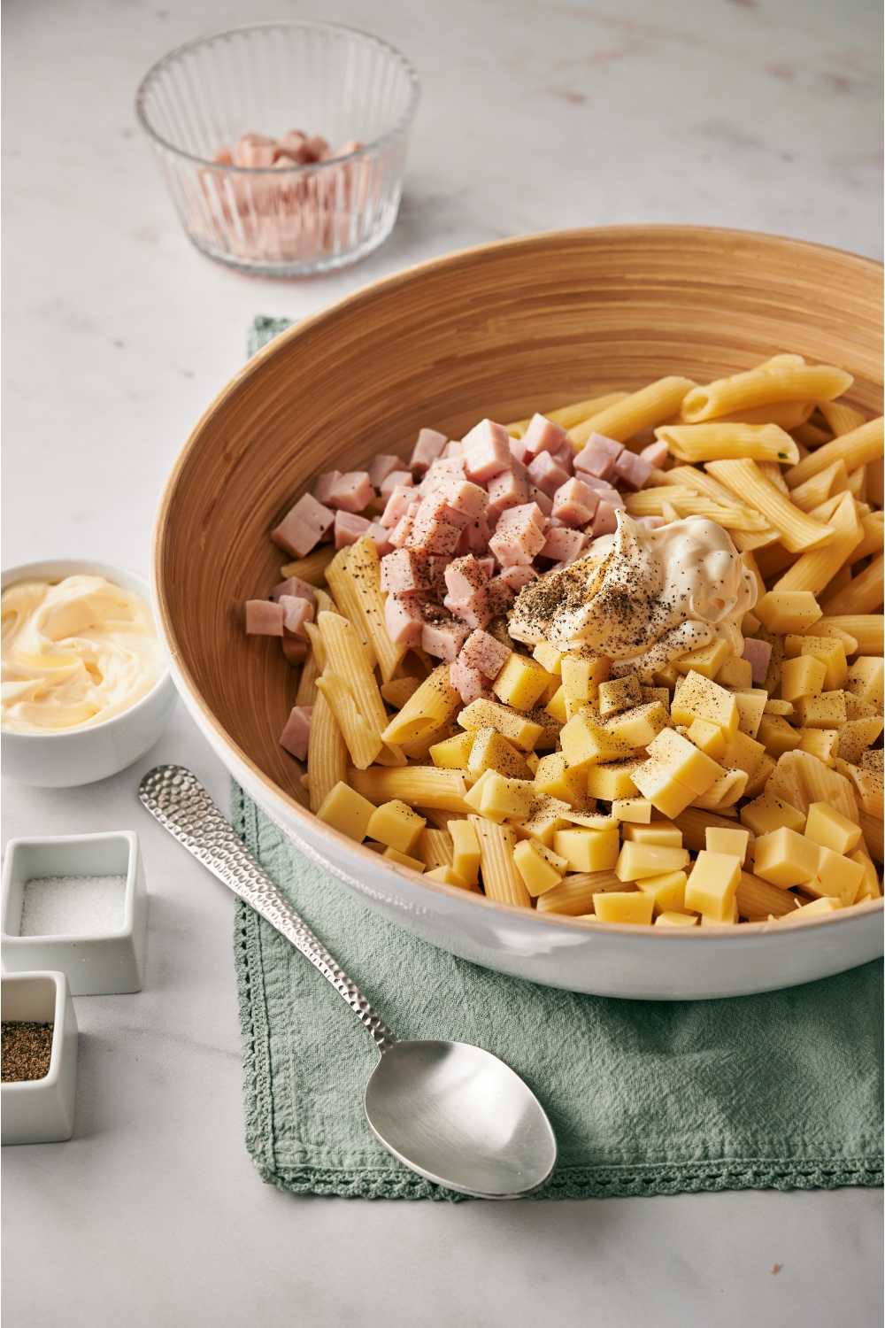 A brown and white bowl with cooked penne pasta, cubed ham, cubed cheese, mayonnaise, and pepper in it, next to a spoon and smaller bowls of mayo and cubed ham.