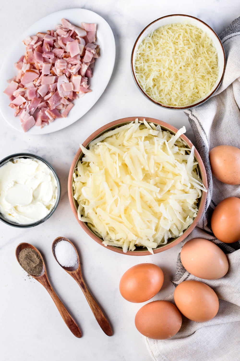 An assortment of ingredients including raw eggs, two spoonfuls of salt and pepper, a plate of diced bacon, and bowls of hash browns, shredded cheese, and cream cheese.