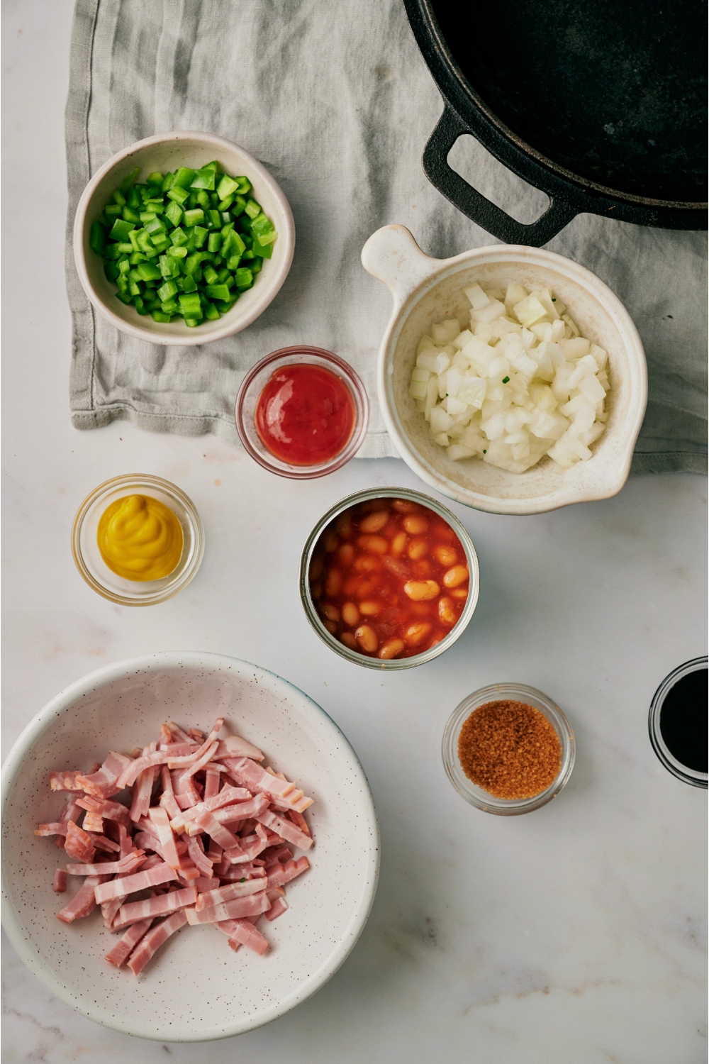 An assortment of ingredients including bowls of raw bacon, ketchup, brown sugar, diced onion, diced green bell pepper, yellow mustard, and an open can of baked beans.