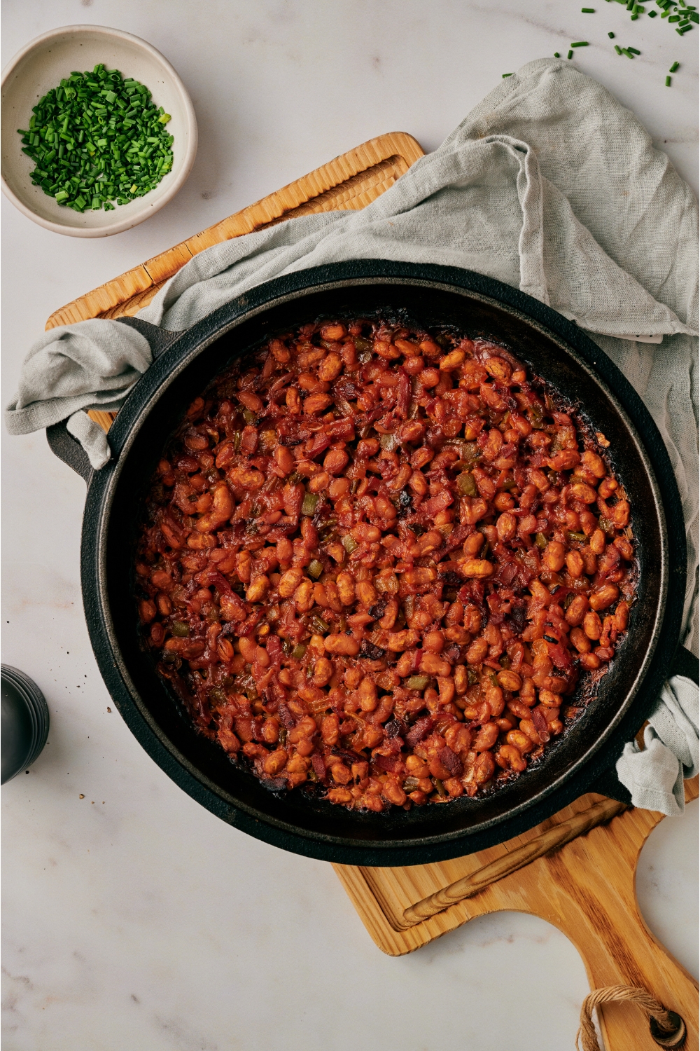 A black skillet atop a wooden board that's filled with freshly baked beans, peppers, and onion.