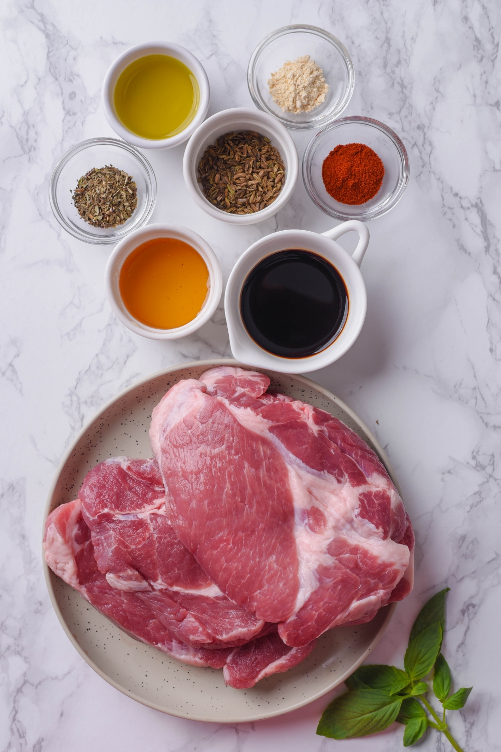 An assortment of ingredients including a plate of raw pork steaks and bowls of soy sauce, oil, spices, and herbs.