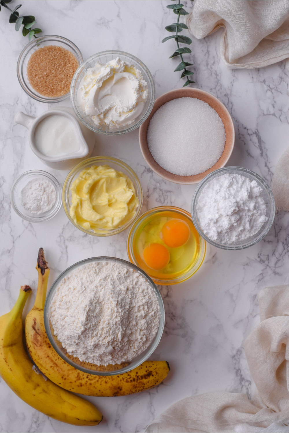 An assortment of ingredients including two bananas and bowls of cream cheese, flour, powdered sugar, white sugar, brown sugar, two eggs, butter, and a pitcher of milk.