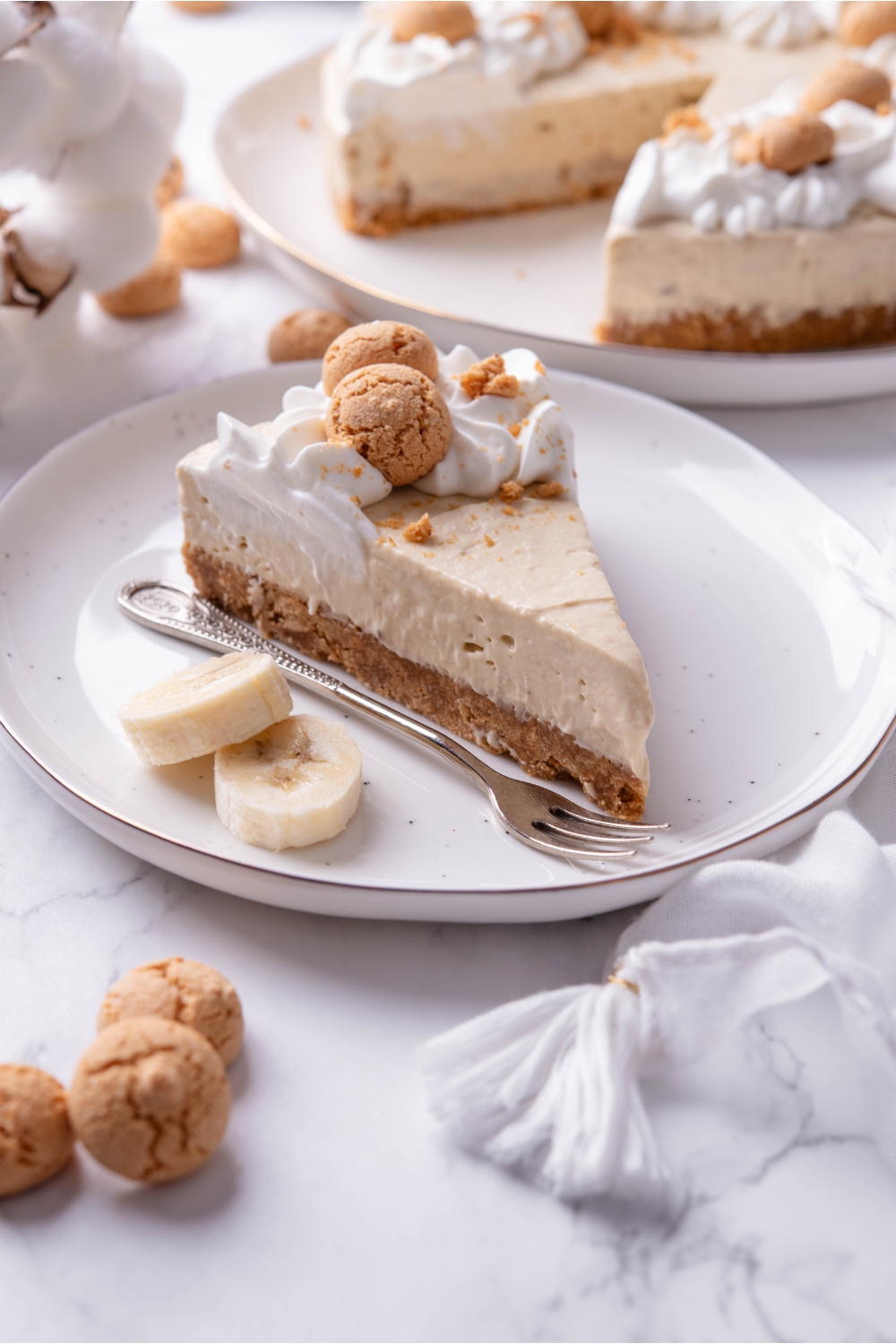 A serving of banana cheesecake on a plate with two banana slices and a fork. The cheesecake has a cookie crust and is topped with whipped cream and mini cookies.