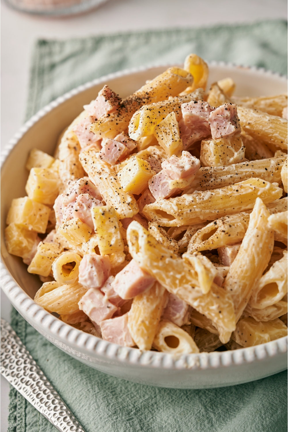 A white bowl filled with pasta salad, with penne pasta, cubed ham, cubed cheese, and black pepper, all tossed in a creamy dressing.