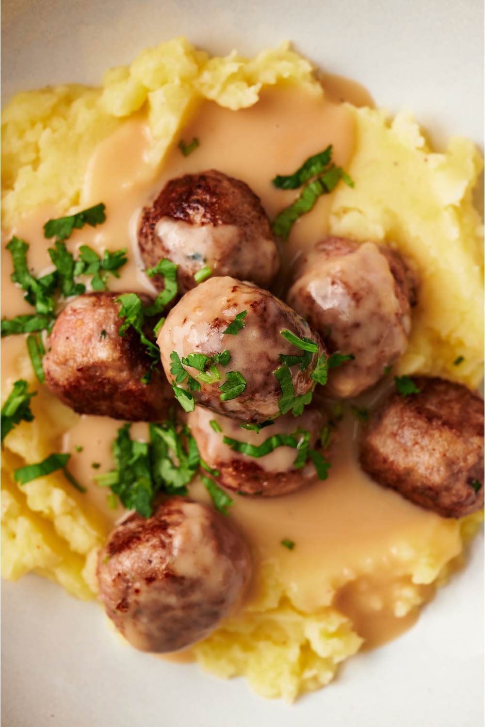 A plate of Swedish meatballs on a bed of mashed potatoes. Creamy gravy covers the potatoes and there is a garnish of fresh green herbs on top.