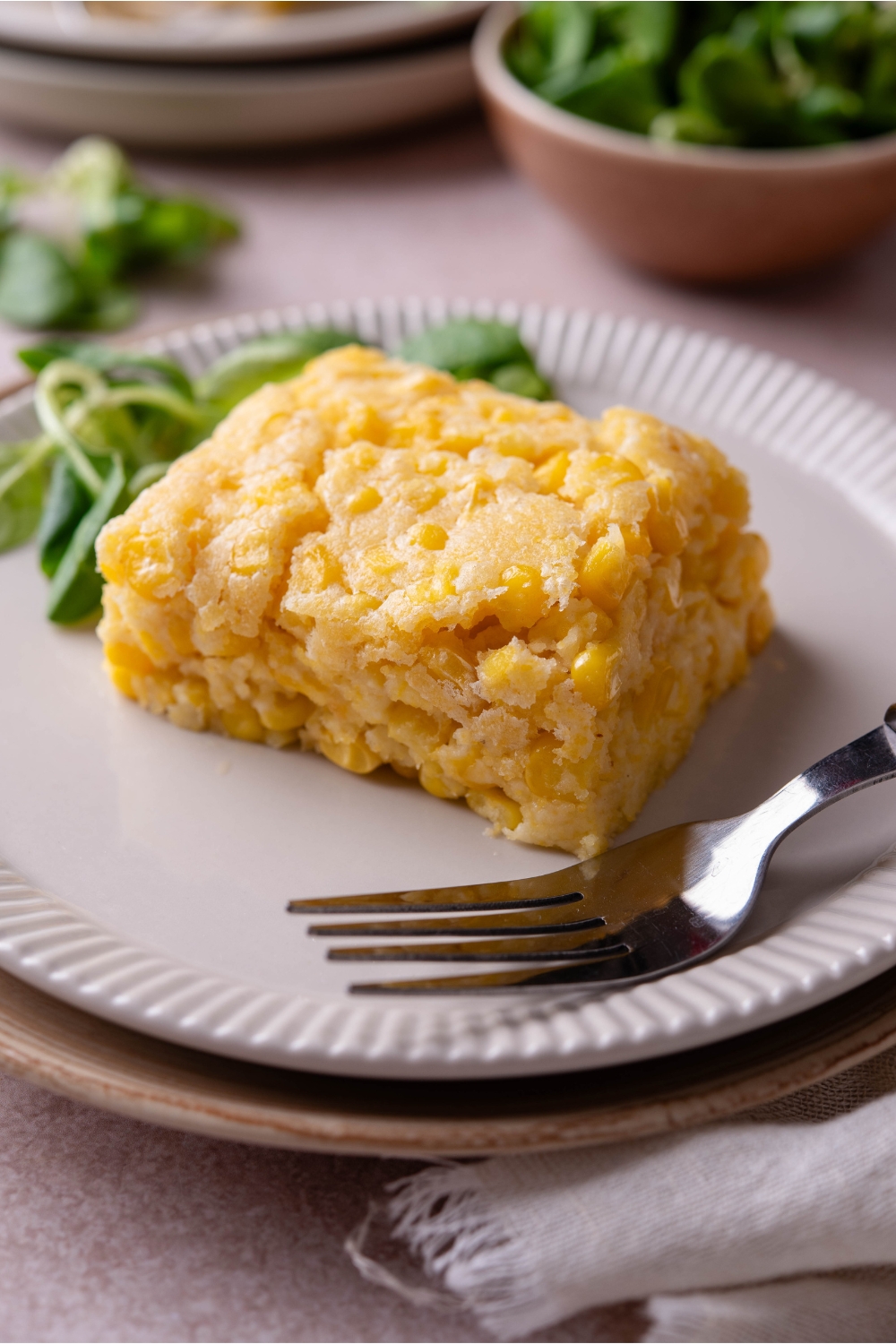 A square of corn casserole with a fork and a pile of fresh greens on the plate.