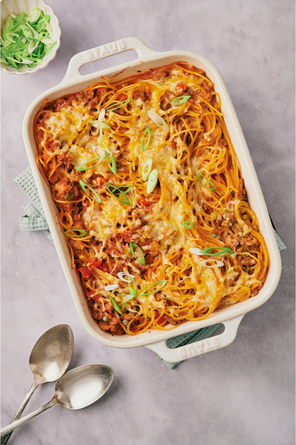 A white baking dish filled with cooked spaghetti casserole covered in a layer of melted cheese and green onions.