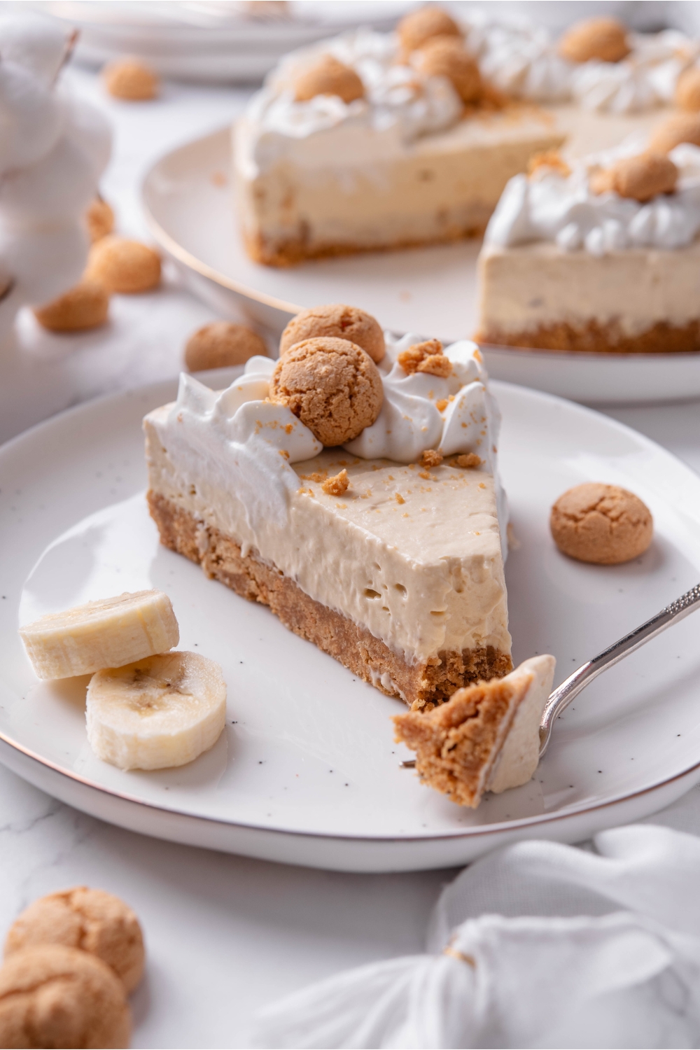 A serving of banana cheesecake on a plate with a bite taken out using a fork that is on the plate. The cheesecake has a cookie crust and is topped with whipped cream and mini cookies.