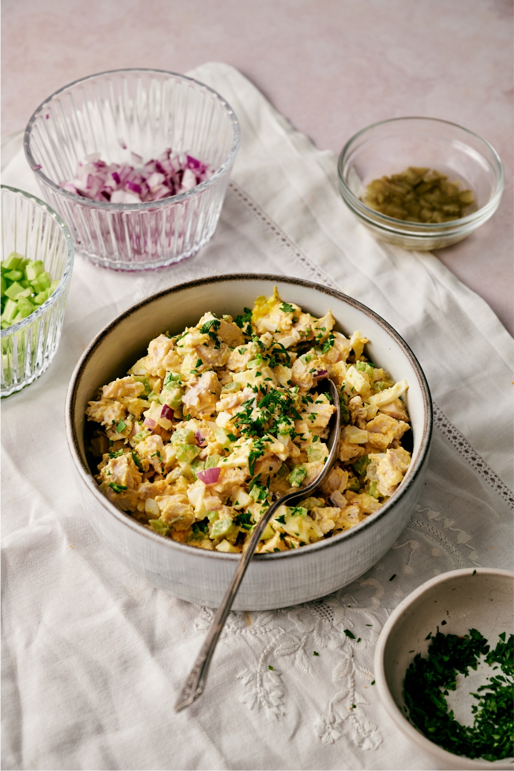 A bowl of chicken and egg salad with pieces of chopped red onion, celery, fresh green herbs, and a spoon in the bowl. The bowl is surrounded by smaller bowls of chopped pickles, red onion, and celery.