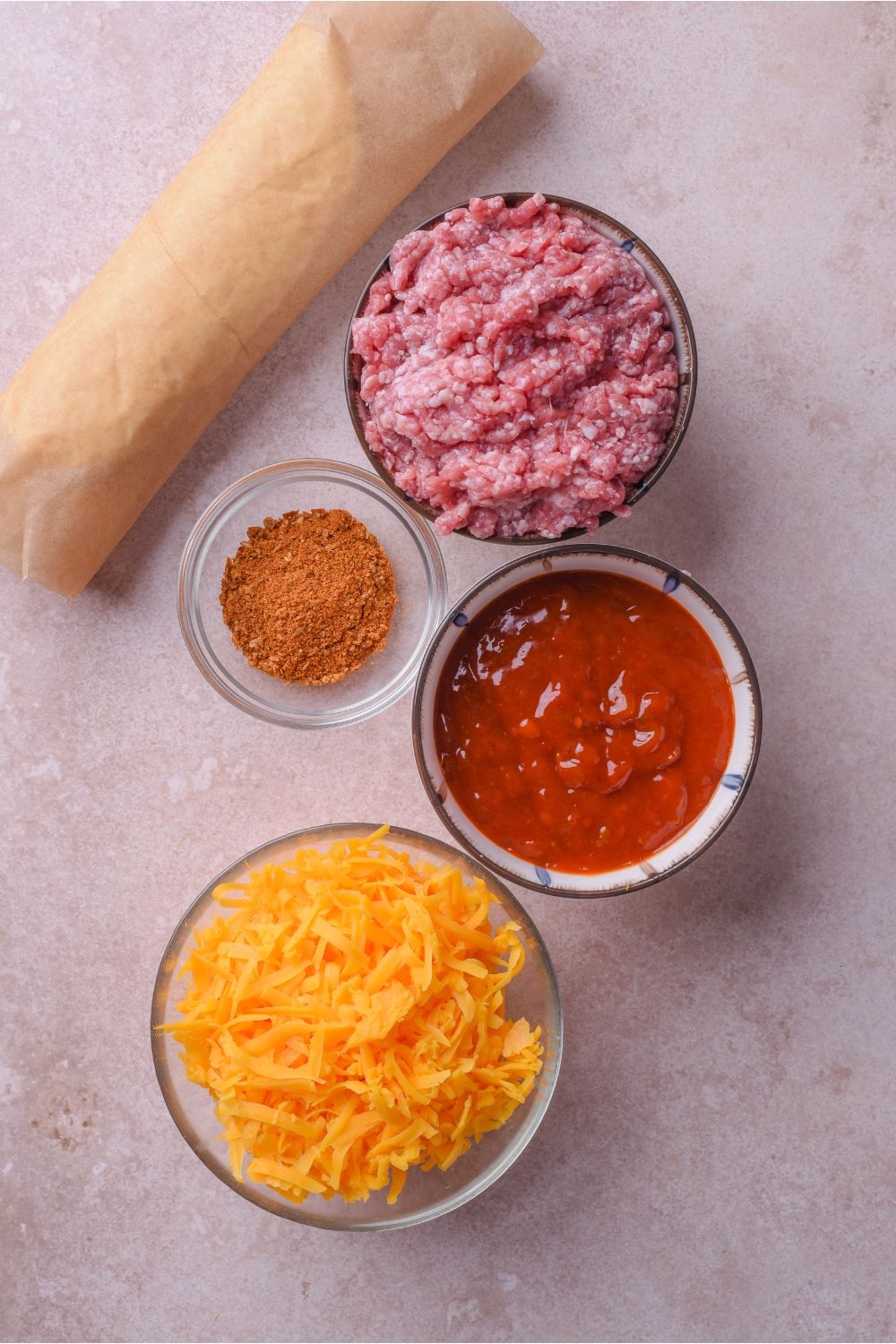 An assortment of ingredients including bowls of salsa, raw ground beef, taco seasoning, shredded cheese, and a roll of dough wrapped in parchment paper.