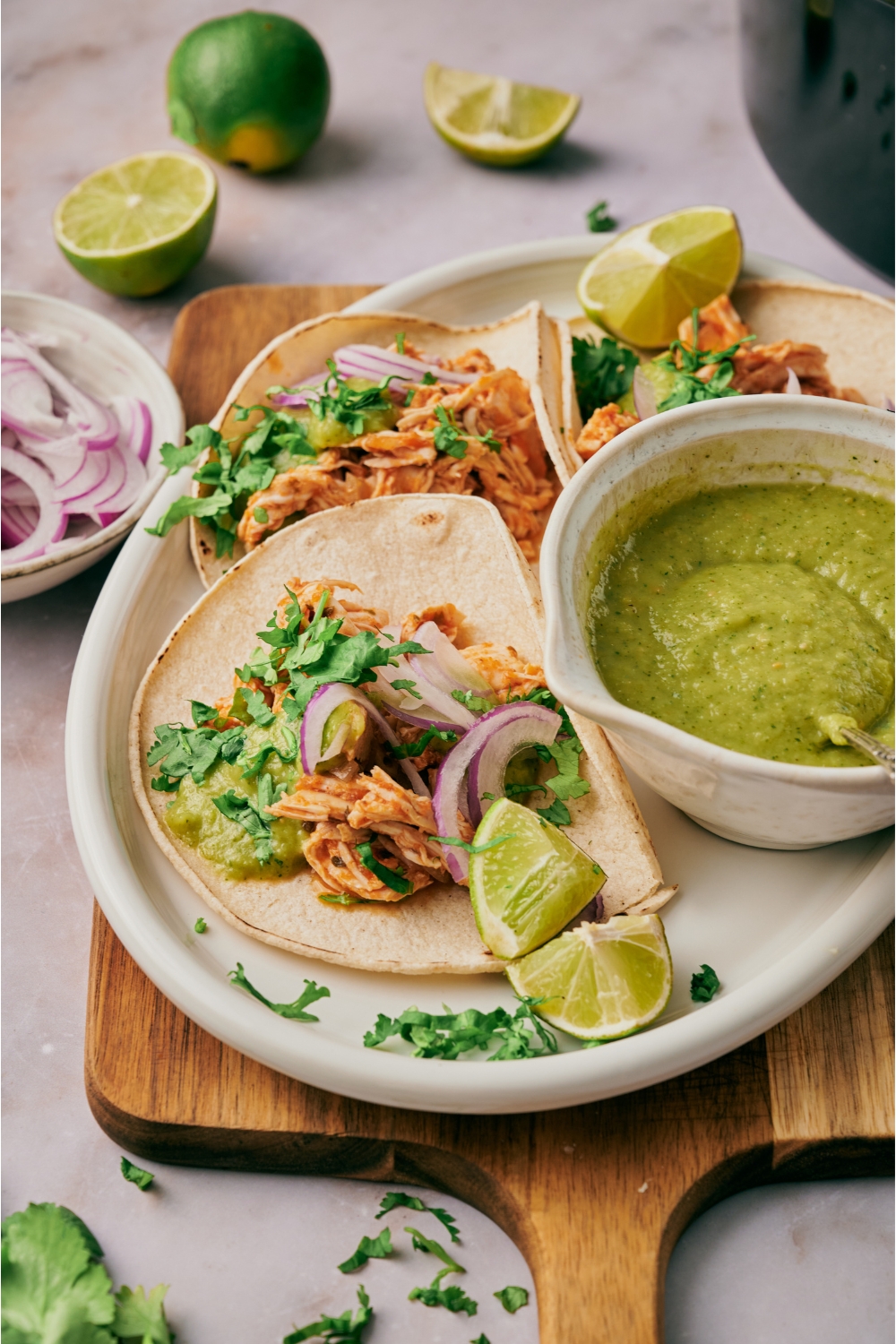 A plate with three shredded chicken tacos topped with sliced red onion and cilantro with a bowl of green salsa and lime wedges on the plate.