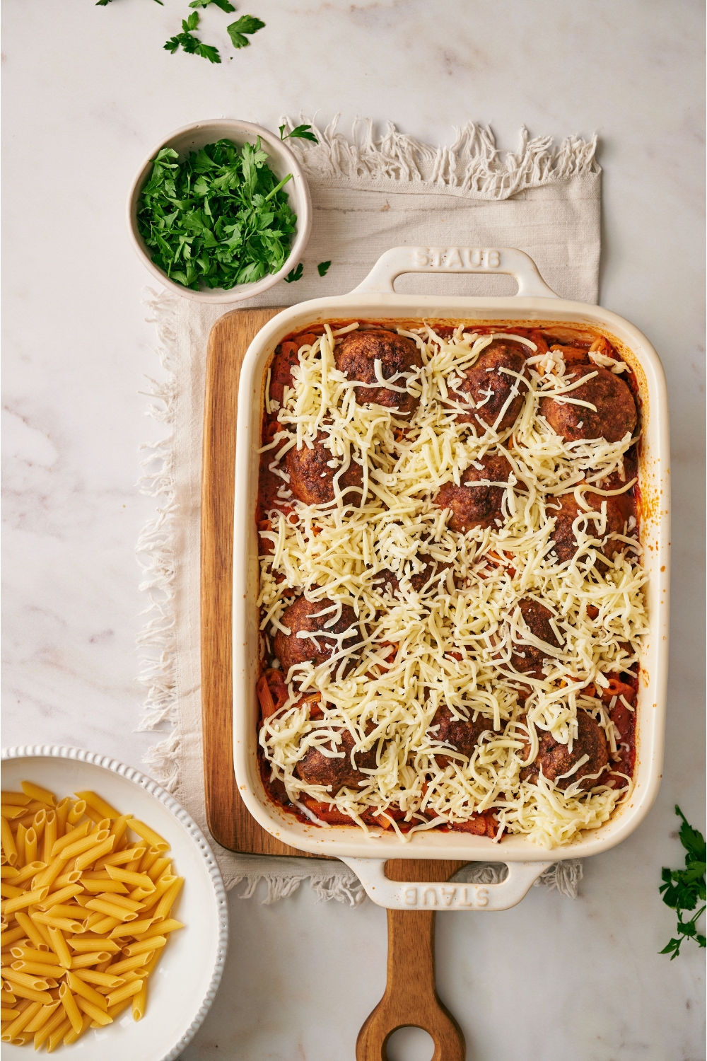 A white baking dish filled with pasta casserole that's been topped with browned meatballs and a layer of shredded cheese.