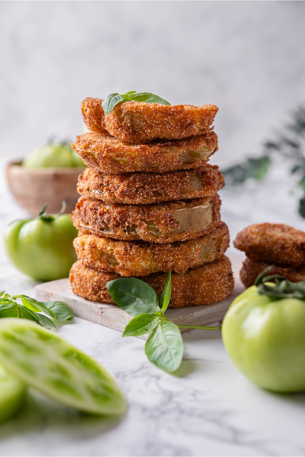 Six breaded and fried green tomatoes stacked evenly on top of each other with a garnish of green herbs on top and fresh green tomatoes surrounding the stack.