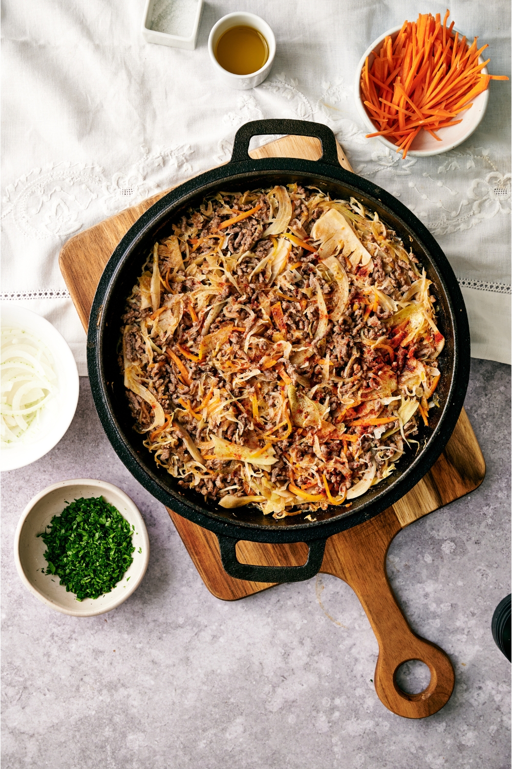 A black skillet filled with ground beef and cabbage stir fry. The skillet is on a wooden board next to bowls of parsley and sliced carrots.