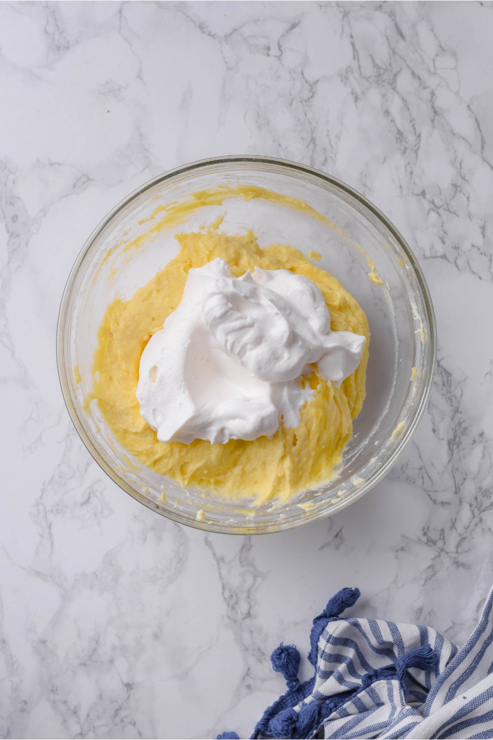 A clear bowl filled with yellow cheesecake batter and a scoop of whipped cream not yet combined.